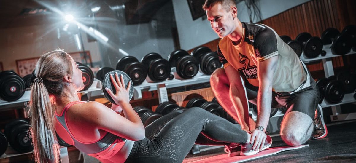 A personal trainer assists a woman doing sit-ups with a medicine ball in a gym setting.